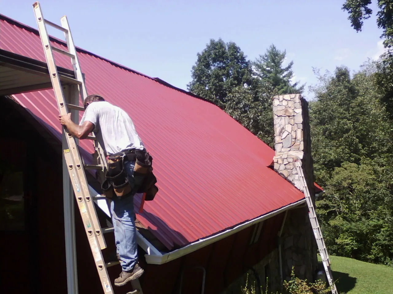 Roofer on a ladder beside a red metal roof and stone chimney — Soffit & Fascia Repair by Roofing Brothers in Anza